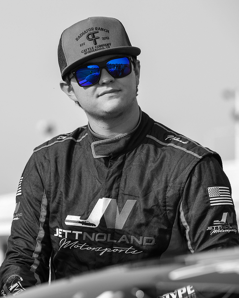 Black and white image of Jett Noland of Jett Noland Motorsports #50 in his race suit and a baseball hat with the only color of in the image being the blue in this sunglass lenses