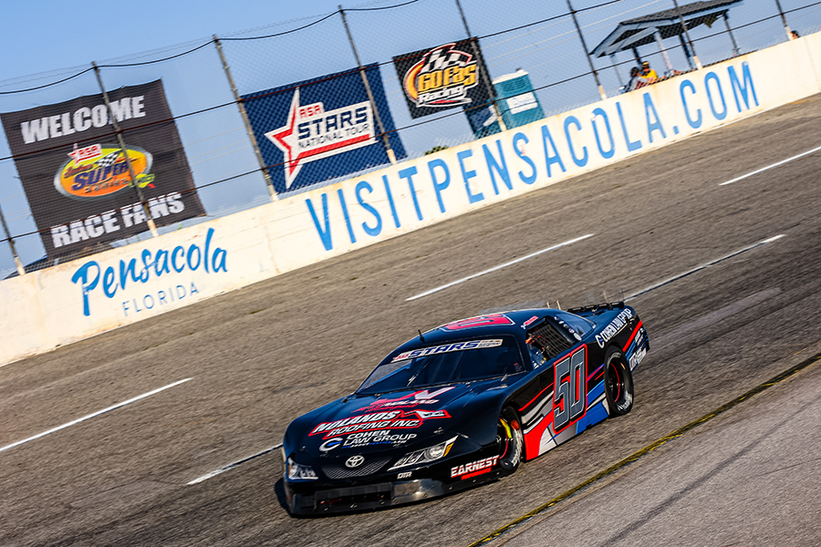 A black stock car, Jett Noland the number 50 and 'Cohen Law Group' sponsorship races on a track at Pensacola, Florida. The car, branded as a Toyota Camry, has a 'ASA STARS' decal on the windshield. The background features banners welcoming race fans and promoting 'VisitPensacola.com'.