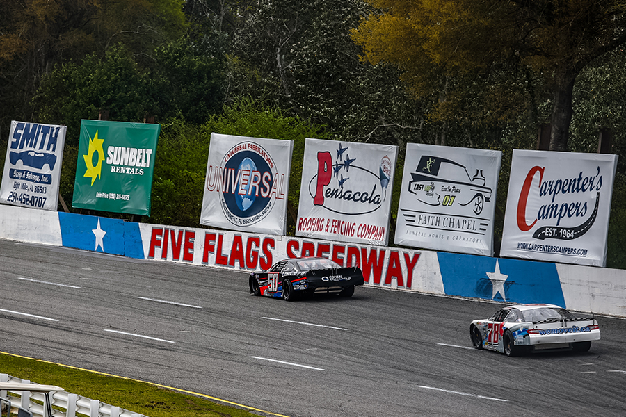 Stock cars racing at Five Flags Speedway in Pensacola, Florida. The black number 50 car, Jett Noland with 'Cohen Law Group' sponsorship and a white number 26 car are seen on the track. The background features banners for various local businesses, including Sunbelt Rentals, Universal, and Carpenter's Campers.
