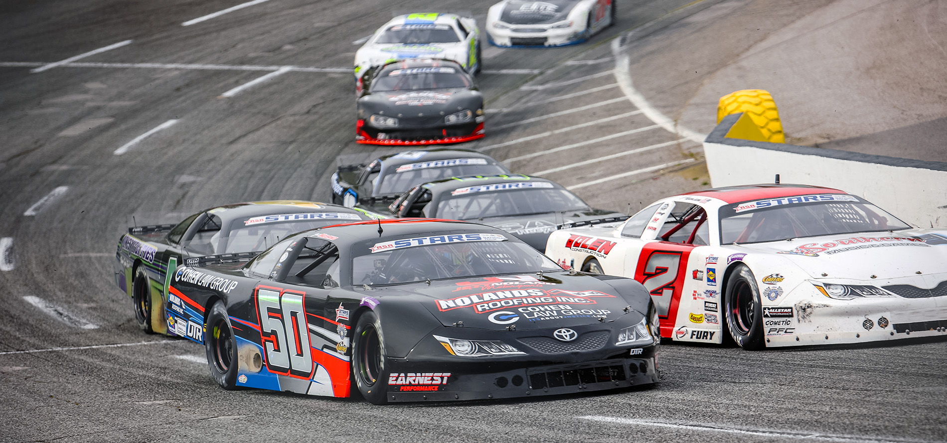 A black stock car with the number 50 Jett Noland with 'Noland's Roofing' and 'Cohen Law Group' sponsorship, branded as a Toyota Camry, races around a curved section of the track. The car navigates close to a white stock car number 2.