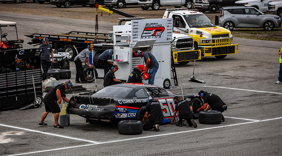 Pit crew performing maintenance on a black race car with the number 50 and Cohen Law Group branding. The team is actively changing tires and refueling the car, with various equipment and support vehicles in the background at a racing event.