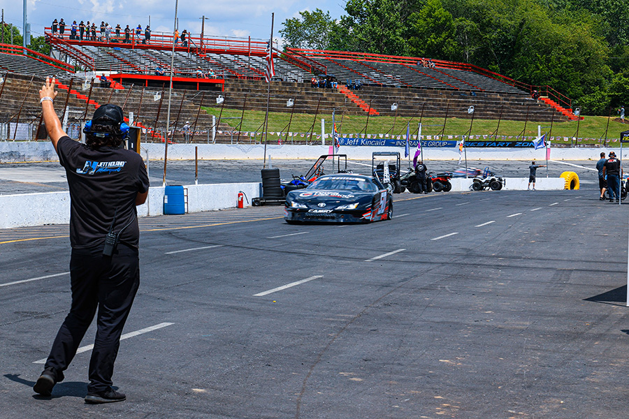 A member of Jett Noland Motorsports pit crew signals to a black stock car with the number 50 as it drives down the pit lane at a racing track. The stands in the background have a few spectators, and there are several team members and equipment along the pit area