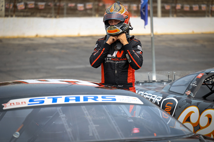 A race car driver, Jett Noland in a black and red racing suit adjusts his helmet while standing next to his vehicle, which has a 'ASA STARS' decal on the windshield. The car is parked on the track, with a white barrier and a few spectators in the background.