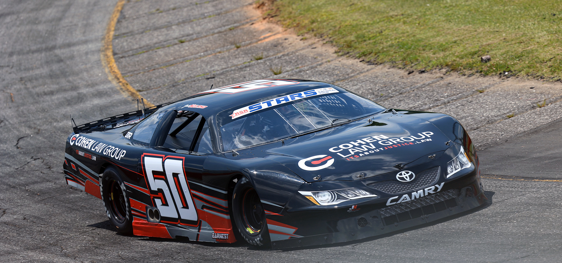A black stock car, Jett Noland's number 50 and 'Cohen Law Group' sponsorship, branded as a Toyota Camry, races around a curved section of the track. The car displays a 'ASA STARS' decal on the windshield and navigates close to the grassy infield.