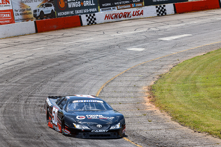 A black stock car with the number 50 - Jett Noland and 'Cohen Law Group' sponsorship races on a curved section of the Hickory,NC USA track. The car navigates close to the grass, with advertisements and a checkered border visible on the barrier in the background.