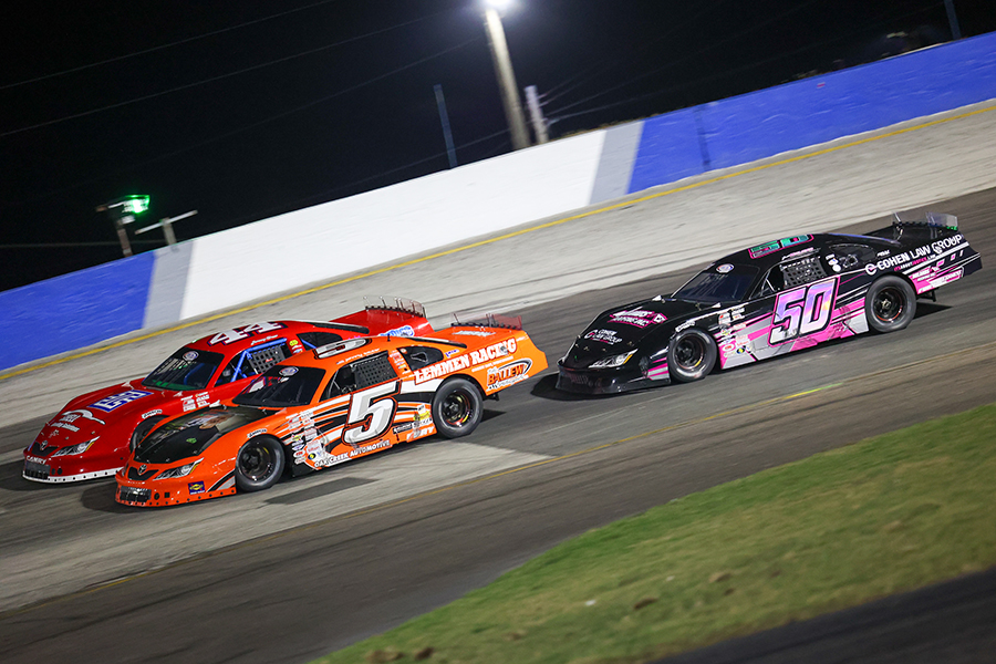 Three race cars competing on the brightly lit track for the Big Rigley Memorial 128 at the Freedom Factory. highlighting the thrilling action of a high-speed motorsport event.