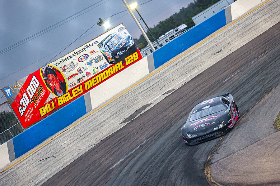 number 50 Jett Noland's race car speeding on a track at the Bill Bigley Memorial 128 event, showcasing a vibrant billboard and the excitement of professional racing.