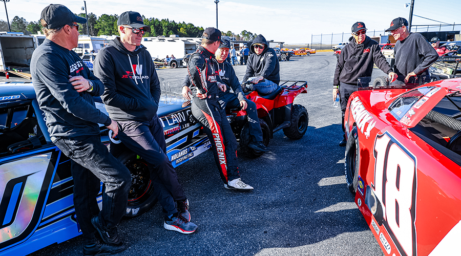 The Jett Noland Motorsports team gathers around their race cars before a race at the track. Team members in branded gear showcase a strong and focused motorsports team atmosphere