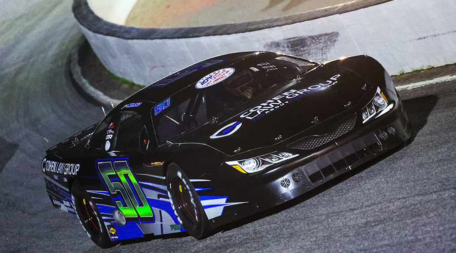 A Jett Noland Motorsports car, with the number 50, speeds around the track at Cordele Motor Speedway during a thrilling night race. The black and blue race car, sponsored by Cohen Law Group, maneuvers through the turn, showcasing precision and speed at the renowned racing venue